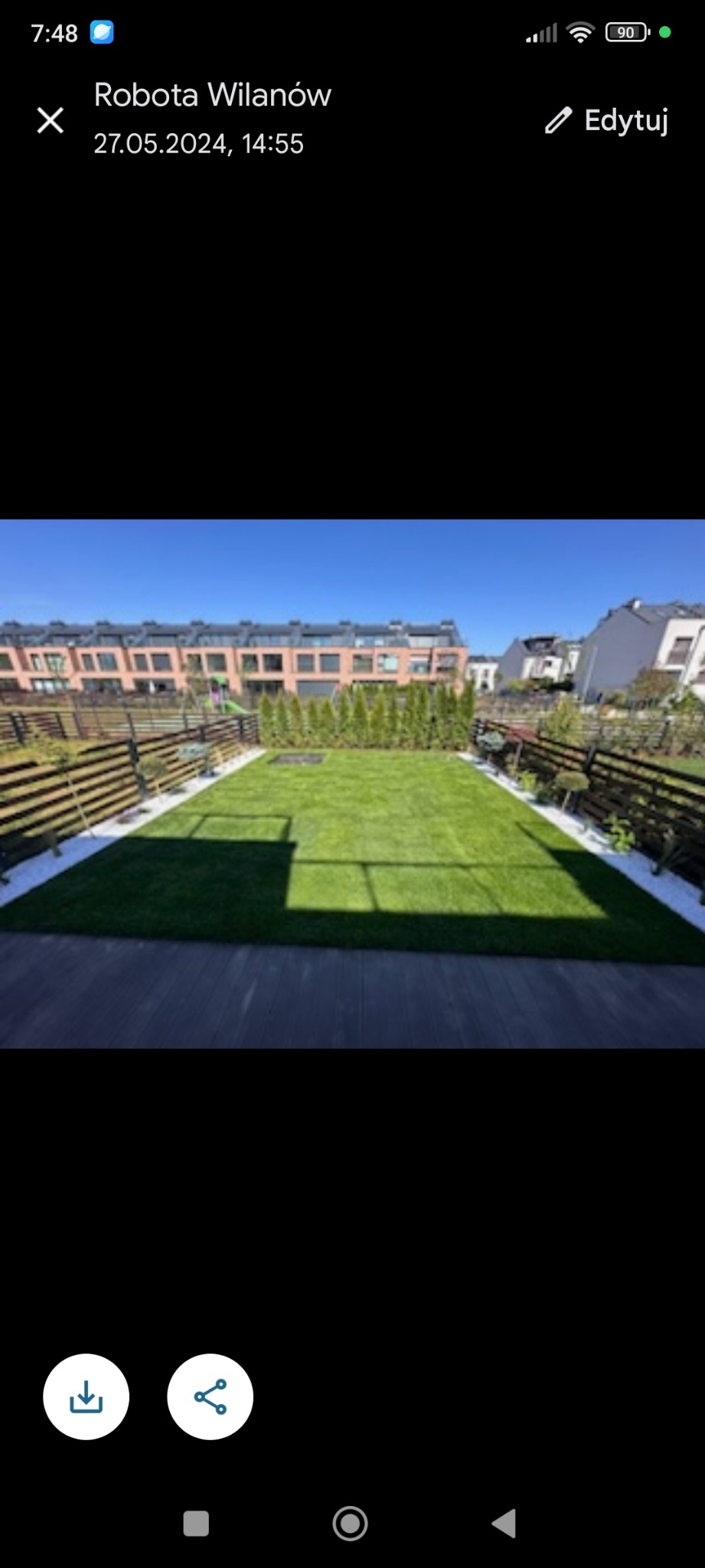 Zadbany trawnik z rolki, obrzeża z białego kamienia, drewniany taras i płot. W tle zabudowa szeregowa w słoneczny dzień. Prace ogrodnicze w Wilanowie.