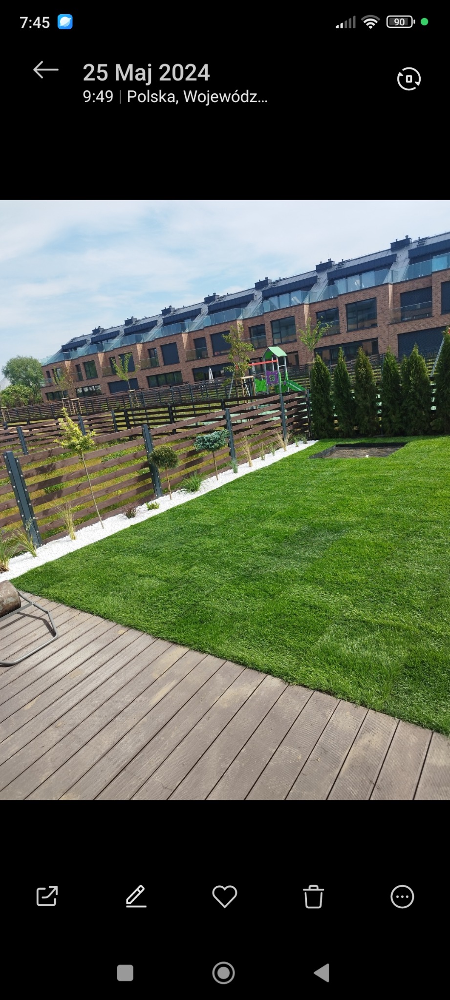 Zadbany trawnik z rolki przylegający do drewnianego tarasu, z widokiem na nowoczesne szeregowce i ogrodzenie z białym kamieniem dekoracyjnym.