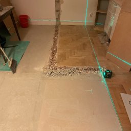 Yhome - Demontaż podłogi w pokoju z widoczną warstwą betonu i fragmentem parkietu. Laserowy niwelator wyznacza poziom. Ściana z odsłoniętą cegłą.