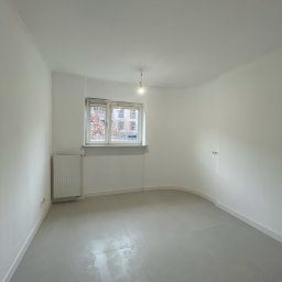 Yhome - Pusty pokój po remoncie: świeżo pomalowane ściany na biało, szara podłoga, grzejnik pod oknem. Widoczne gniazdka elektryczne na ścianie i żarówka na kablu.