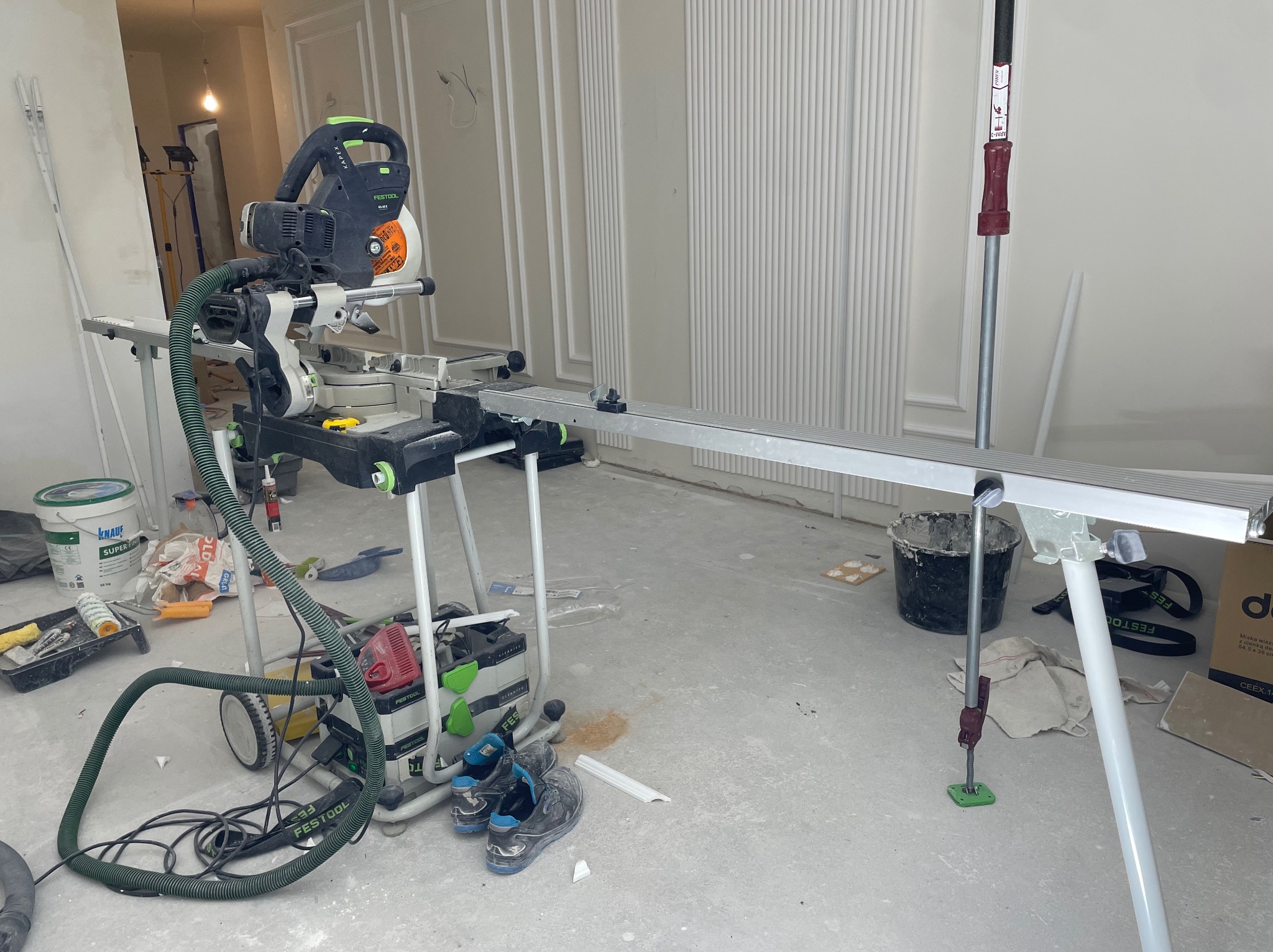 Piła ukośnica Festool na tle ściany z dekoracyjnymi listwami. Widoczne narzędzia i materiały budowlane, buty robocze. Wnętrze w trakcie wykańczania.