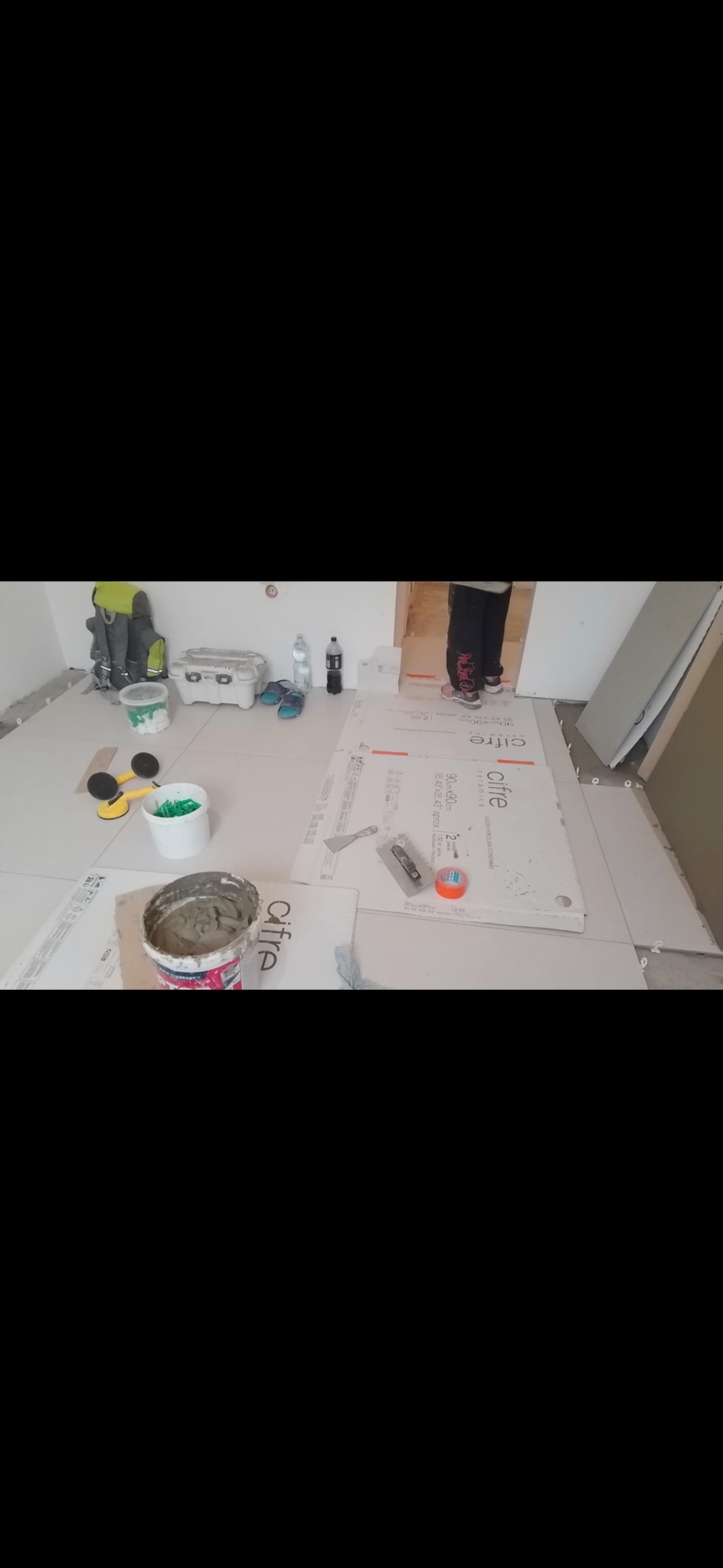 Układanie płytek podłogowych Cifre, widoczne narzędzia i materiały: wiaderko z klejem, paca, poziomnica, przyssawki. Częściowo widoczna osoba w trakcie pracy.