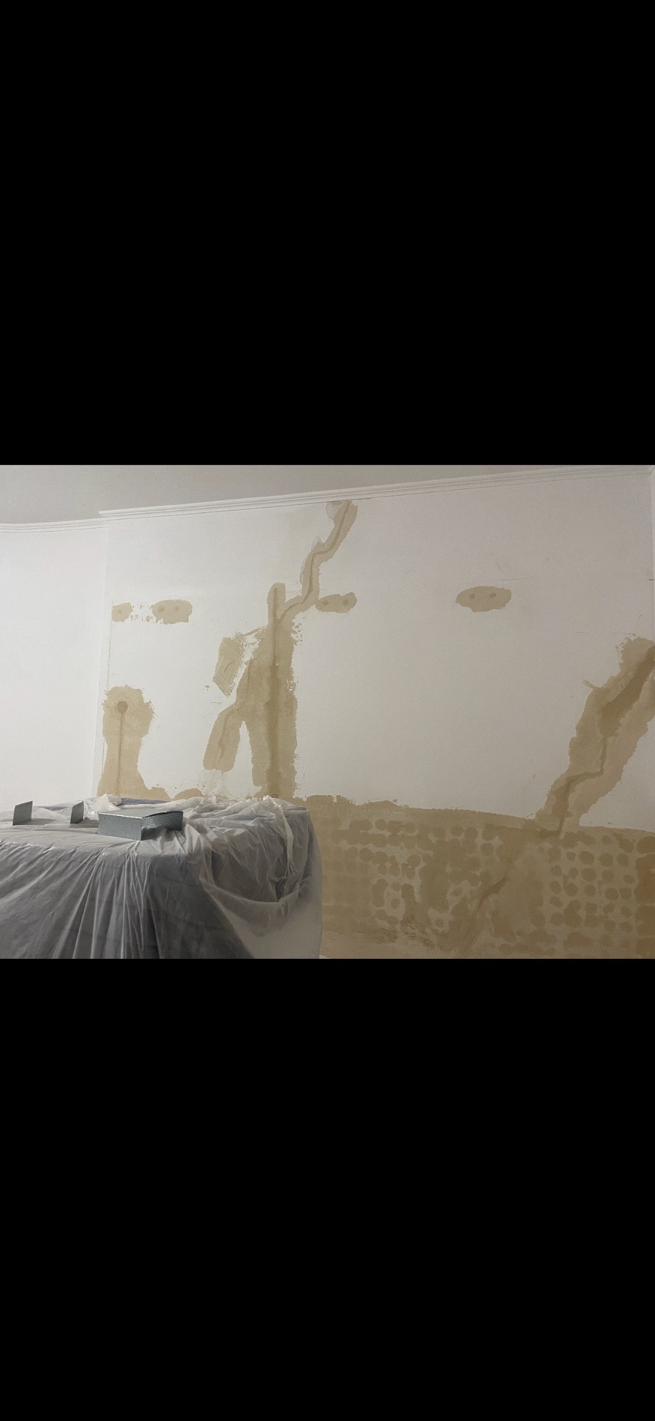 Ściana w trakcie szpachlowania z widocznymi warstwami gładzi, zabezpieczone meble folią malarską. Widok na fragment pokoju w trakcie prac przygotowawczych.