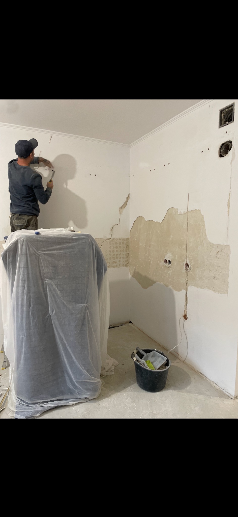 Pracownik w trakcie skuwania tynku w łazience. Widoczne narzędzia, odsłonięta instalacja elektryczna i zabezpieczone meble folią malarską. Fragment ściany bez tynku.