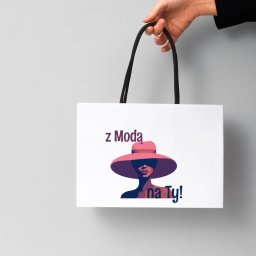 Katarzyna Janek - Biała torba z czarnymi uchwytami, na której widnieje grafika kobiety w kapeluszu i napis 'z Modą na Ty!', trzymana w dłoni na tle szarego tła.