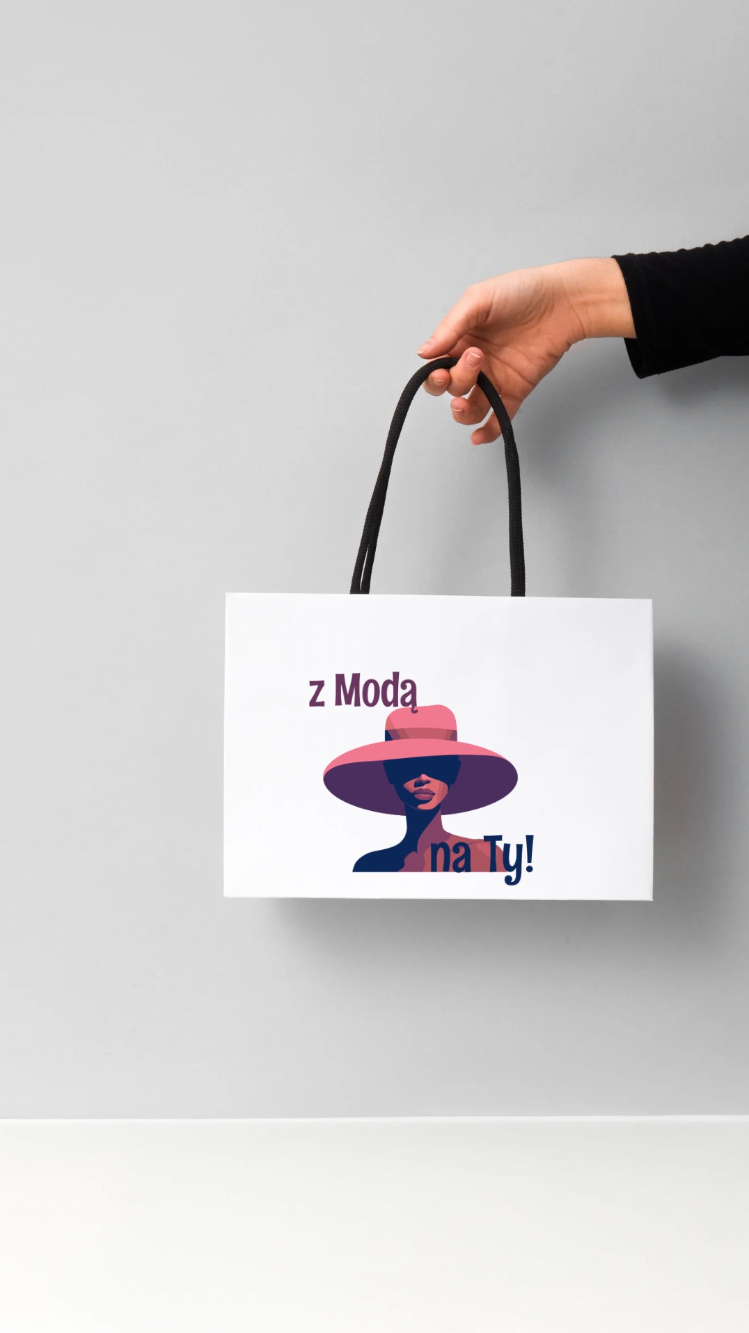 Biała torba z czarnymi uchwytami, na której widnieje grafika kobiety w kapeluszu i napis 'z Modą na Ty!', trzymana w dłoni na tle szarego tła.