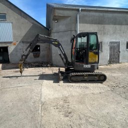 MAREK MACIOSZEK MARKMIT MAREK MACIOSZEK - Czarno-żółta minikoparka Volvo ECR25D z wiertłem hydraulicznym na betonowym placu, na tle szarego budynku z niebieskim niebem w tle.