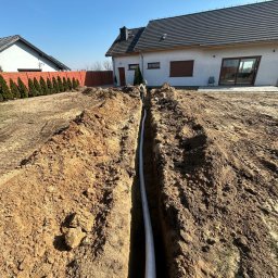 MAREK MACIOSZEK MARKMIT MAREK MACIOSZEK - Wykop z białą rurą w ziemi, na tle domu jednorodzinnego i ogrodzenia z tujami. Widoczny pracownik w oddali. Wykop świeży, ziemia brązowa.