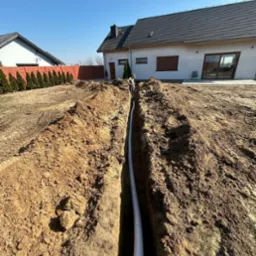 Wykop z białą rurą w ziemi, na tle domu jednorodzinnego i ogrodzenia z tujami. Widoczny pracownik w oddali. Wykop świeży, ziemia brązowa.