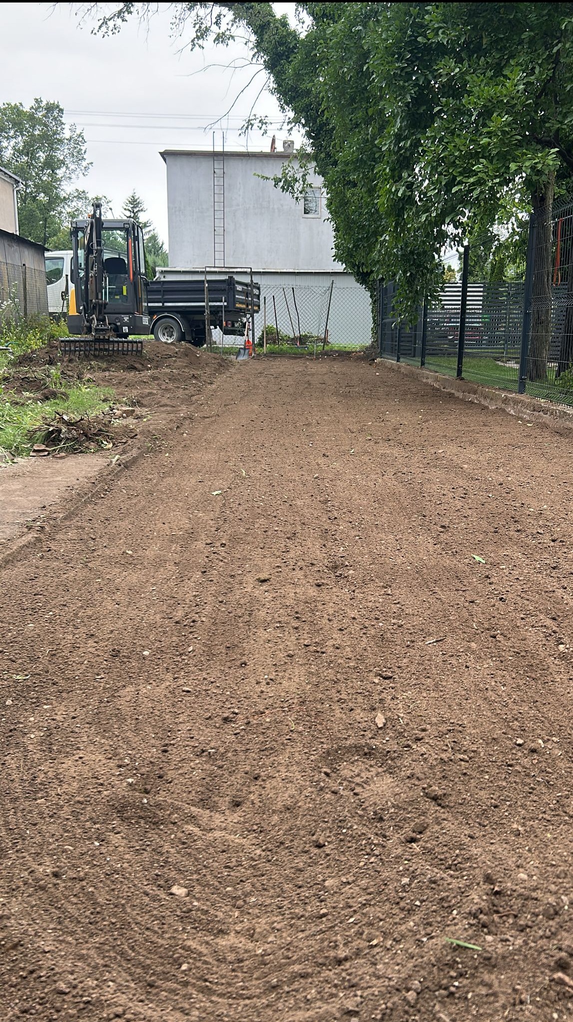 Wyrównany teren z mini wywrotką i grabiami w tle. Przygotowany grunt pod trawnik lub ogród, ogrodzenie siatkowe z prawej strony.