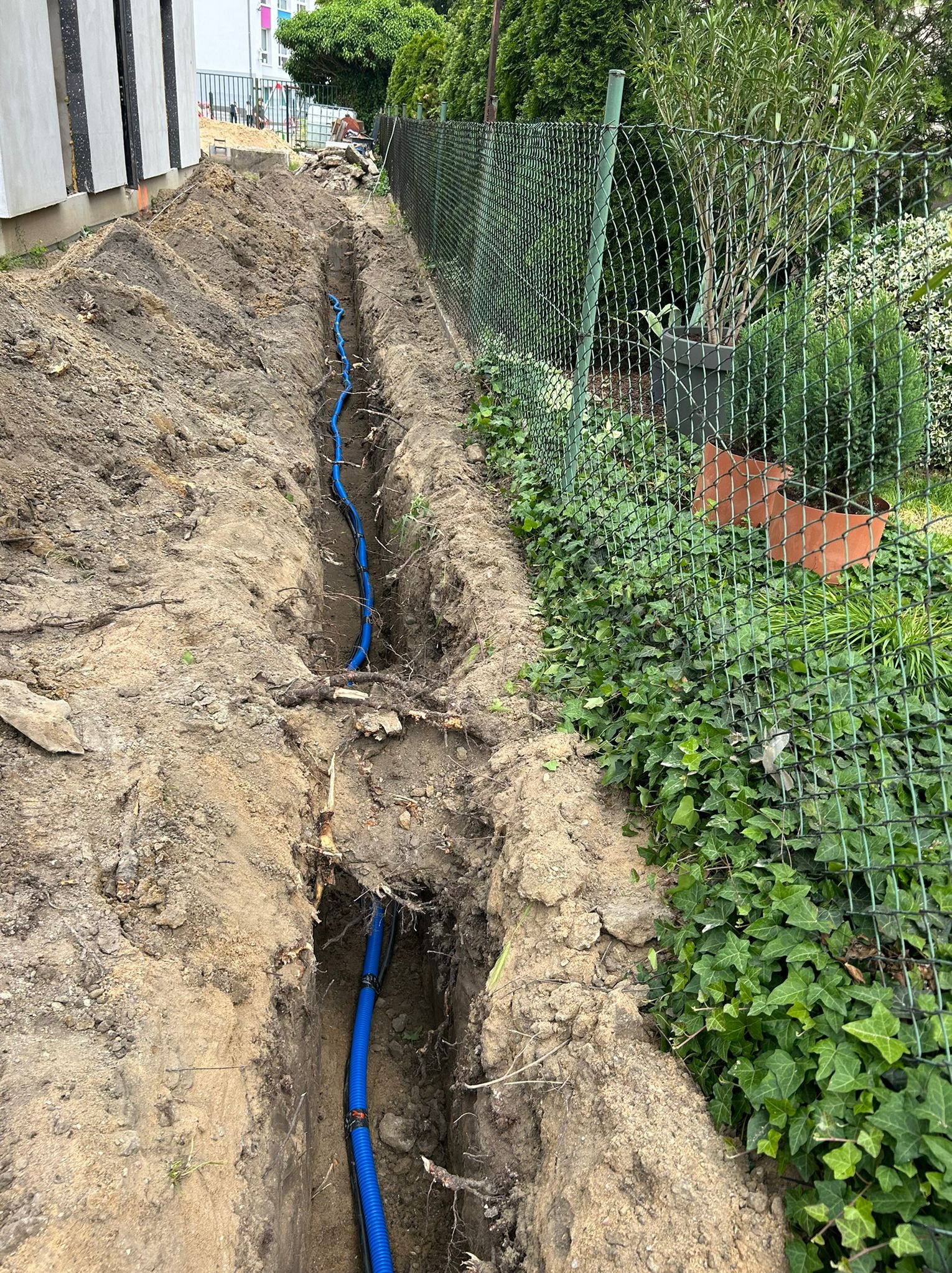 Wykop z niebieskim przewodem w ziemi, widok z góry. Prace ziemne przy ogrodzeniu z siatki. Wykopana ziemia po bokach. Etap instalacji przyłącza wod-kan.