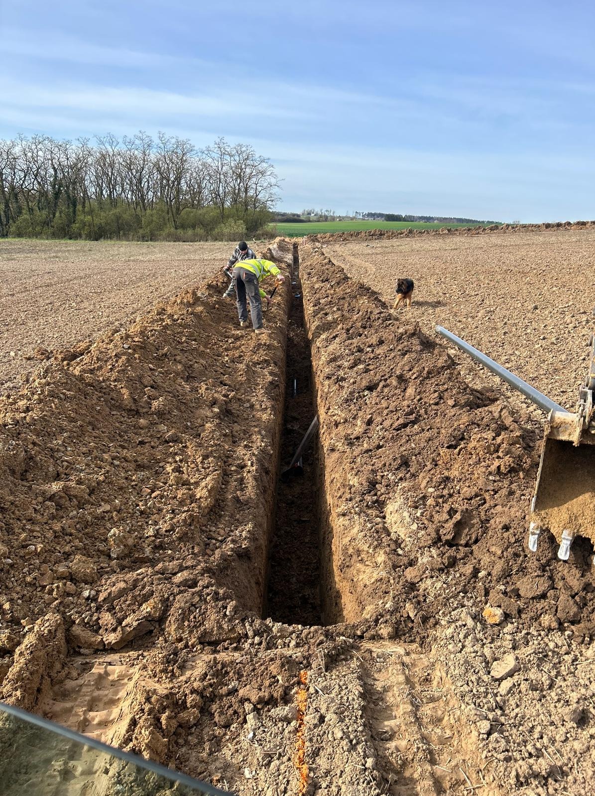 Wykopany głęboki rów w polu, dwaj robotnicy w odblaskowych kamizelkach pracują w środku, obok stoi pies i fragment koparki. Przygotowanie do instalacji.