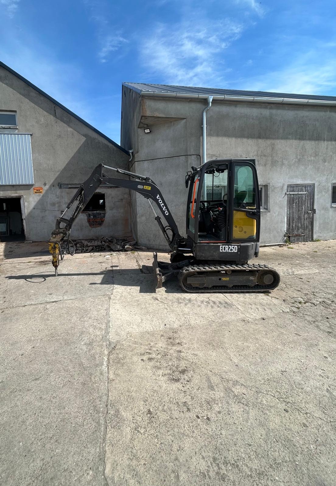Czarno-żółta minikoparka Volvo ECR25D z wiertłem hydraulicznym na betonowym placu, na tle szarego budynku z niebieskim niebem w tle.