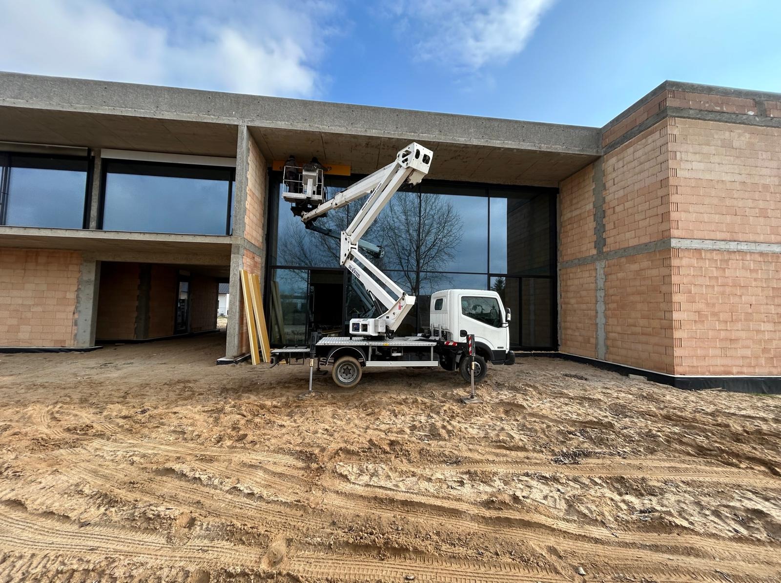 Podnośnik koszowy na tle surowego budynku z dużymi oknami. Biała maszyna z wysięgnikiem w trakcie prac na wysokości. Teren wokół budynku piaszczysty.