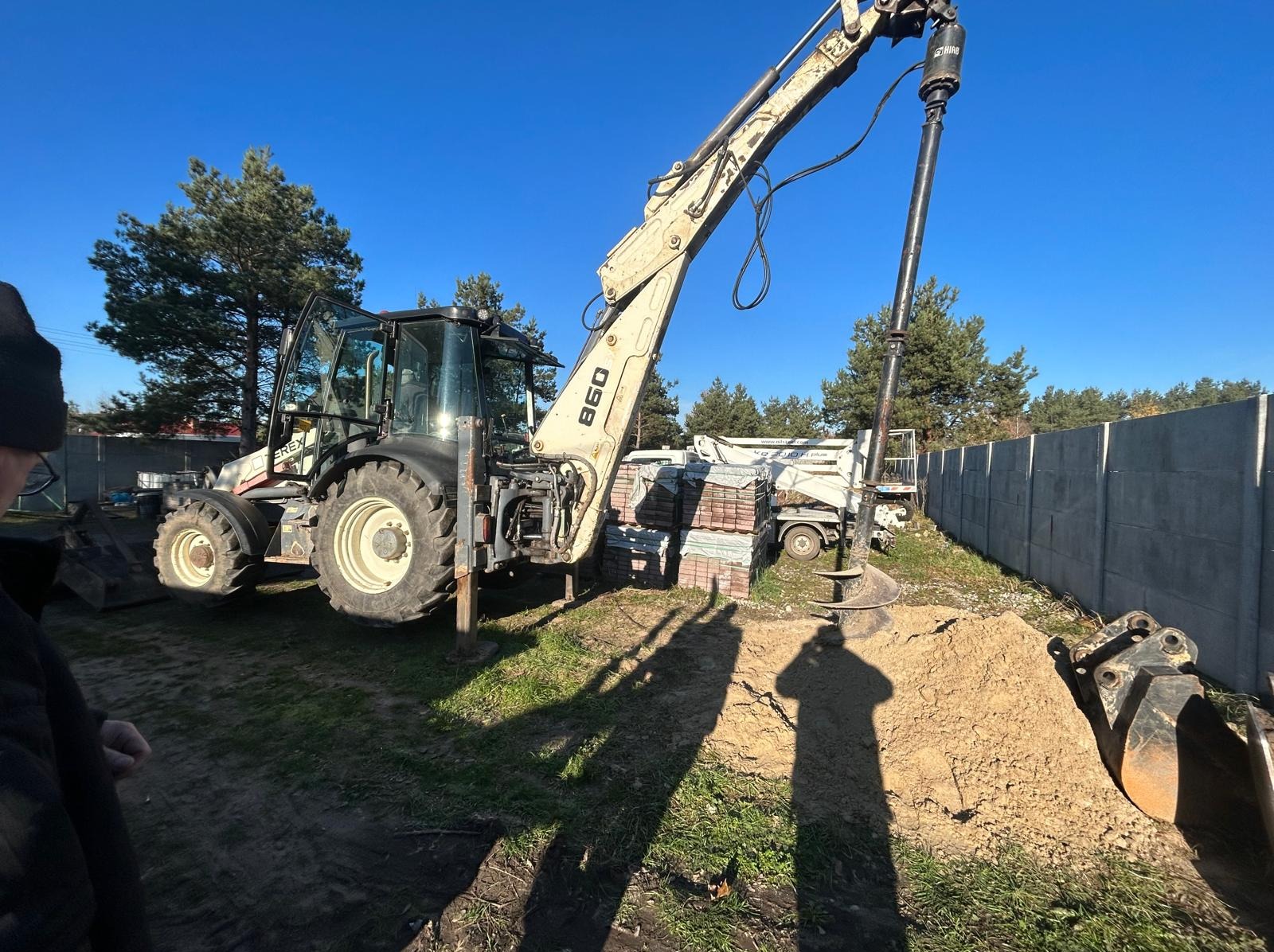 Wiertnica glebowa zamontowana na koparko-ładowarce w Kostrzynie, wierci otwór w ziemi. W tle palety z kostką brukową i betonowe ogrodzenie.