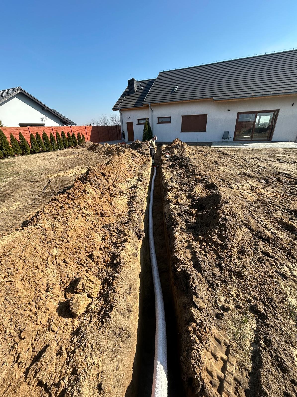 Wykop z białą rurą w ziemi, na tle domu jednorodzinnego i ogrodzenia z tujami. Widoczny pracownik w oddali. Wykop świeży, ziemia brązowa.