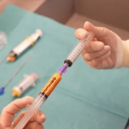 Przygotowanie do iniekcji komórek macierzystych: strzykawka z pomarańczowym płynem w dłoni w rękawiczce, inne strzykawki i narzędzia na zielonym podkładzie.