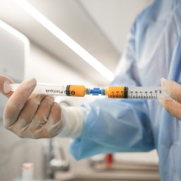 ArthroClinic Ortopedia i Fizjoterapia Puck - Dwie strzykawki połączone łącznikiem, wypełnione pomarańczowym płynem, trzymane w dłoniach w rękawiczkach, na tle jasnego, sterylnego pomieszczenia zabiegowego.