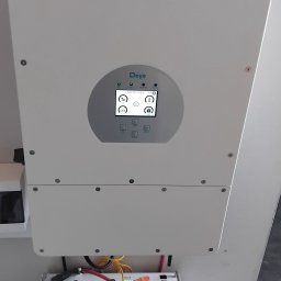 SOLAR SYSTEM Halina Karoń - Biały magazyn energii Deye z ekranem, z widocznymi modułami bateryjnymi i przewodami, zamontowany na ścianie. Widoczne gniazdka elektryczne.