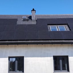 SOLAR SYSTEM Halina Karoń - Czarny panel słoneczny na szarym dachu domu z dwoma oknami dachowymi i kominem na tle błękitnego nieba. Widok z dołu na elewację budynku.