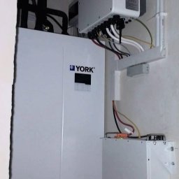 SOLAR SYSTEM Halina Karoń - Instalacja wewnętrzna: biały moduł YORK z czarnymi rurami, nad nim falownik z wyświetlaczem i okablowaniem, a poniżej moduł Deye. Białe ściany, surowy stan.