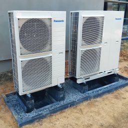 SOLAR SYSTEM Halina Karoń - Dwie jednostki zewnętrzne pompy ciepła Panasonic, zamontowane na betonowych podstawach z kamiennym wypełnieniem, na tle budynku w budowie.