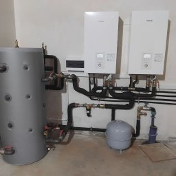 SOLAR SYSTEM Halina Karoń - Szary zbiornik akumulacyjny z rurami i dwa białe moduły Panasonic na ścianie, instalacja grzewcza w kotłowni, widok z przodu, oświetlenie górne.
