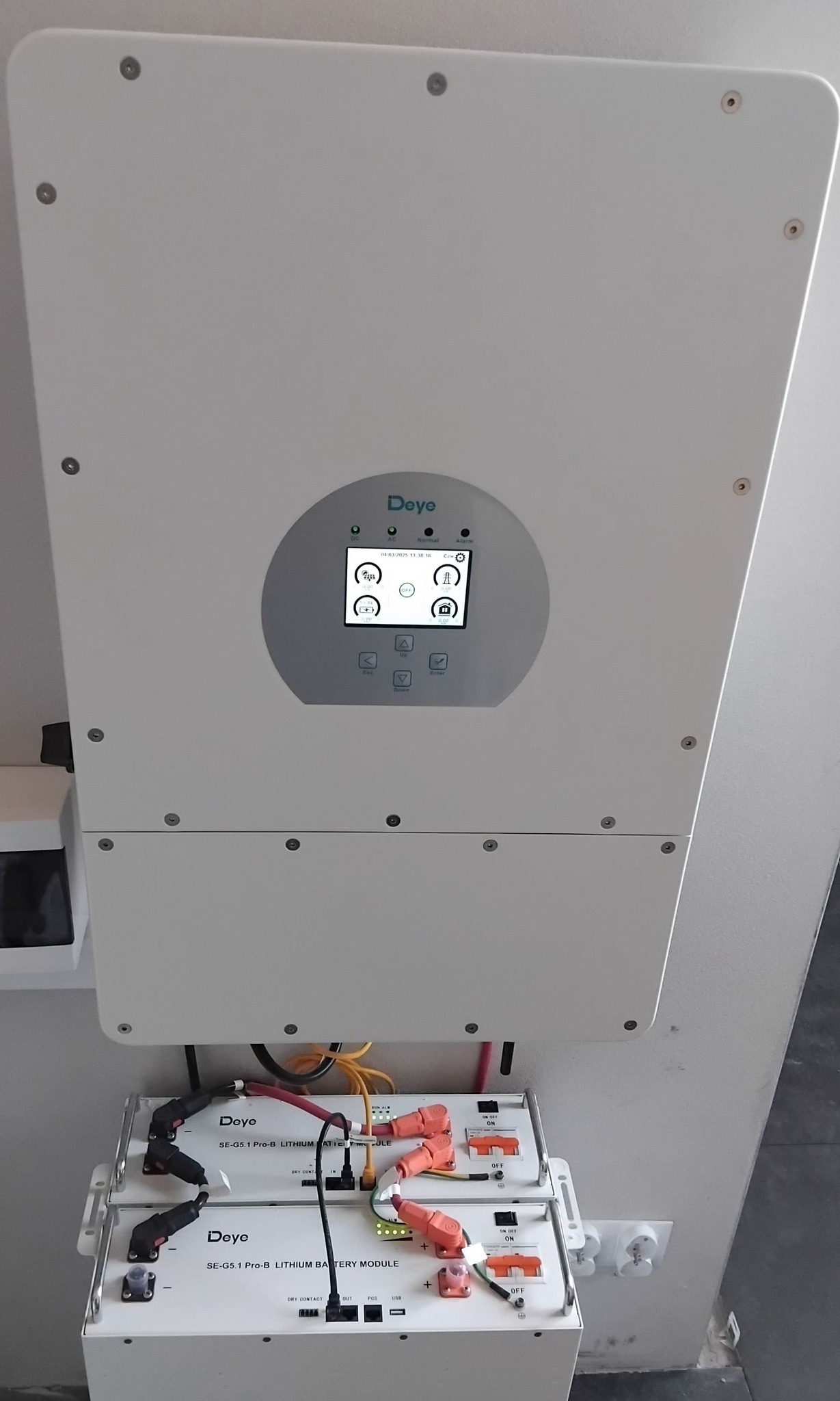 Biały magazyn energii Deye z ekranem, z widocznymi modułami bateryjnymi i przewodami, zamontowany na ścianie. Widoczne gniazdka elektryczne.