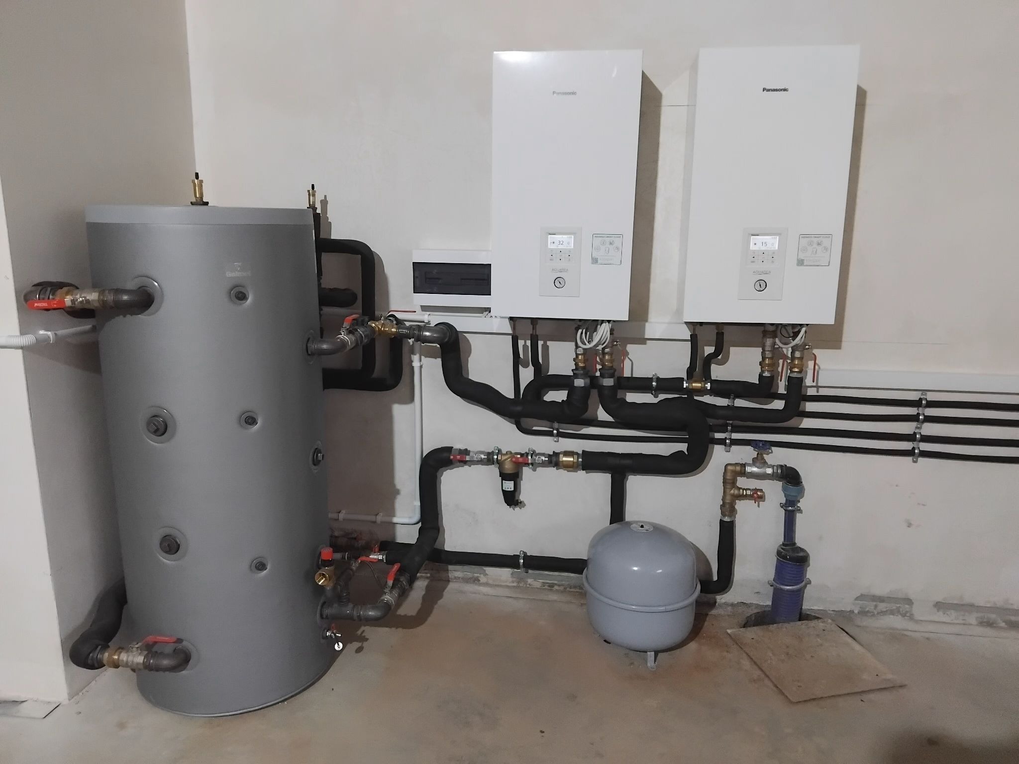 Szary zbiornik akumulacyjny z rurami i dwa białe moduły Panasonic na ścianie, instalacja grzewcza w kotłowni, widok z przodu, oświetlenie górne.