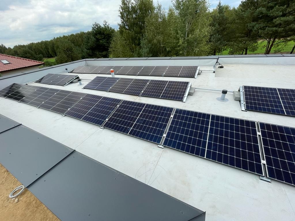 Instalacja fotowoltaiczna na płaskim dachu budynku jednorodzinnego, widok z góry. Moduły w rzędach, szare obróbki blacharskie, zielone drzewa w tle.