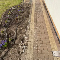 Kaskadowy układ kostki brukowej z żółtymi pasami, obok rabata z kwiatami i systemem nawadniania. Widok z góry na ścieżkę przy budynku.