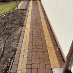 Ułożona, dwukolorowa kostka brukowa (brąz i beż) tworząca ścieżkę wzdłuż białej ściany budynku. Widoczny system nawadniania wzdłuż rabaty kwiatowej.