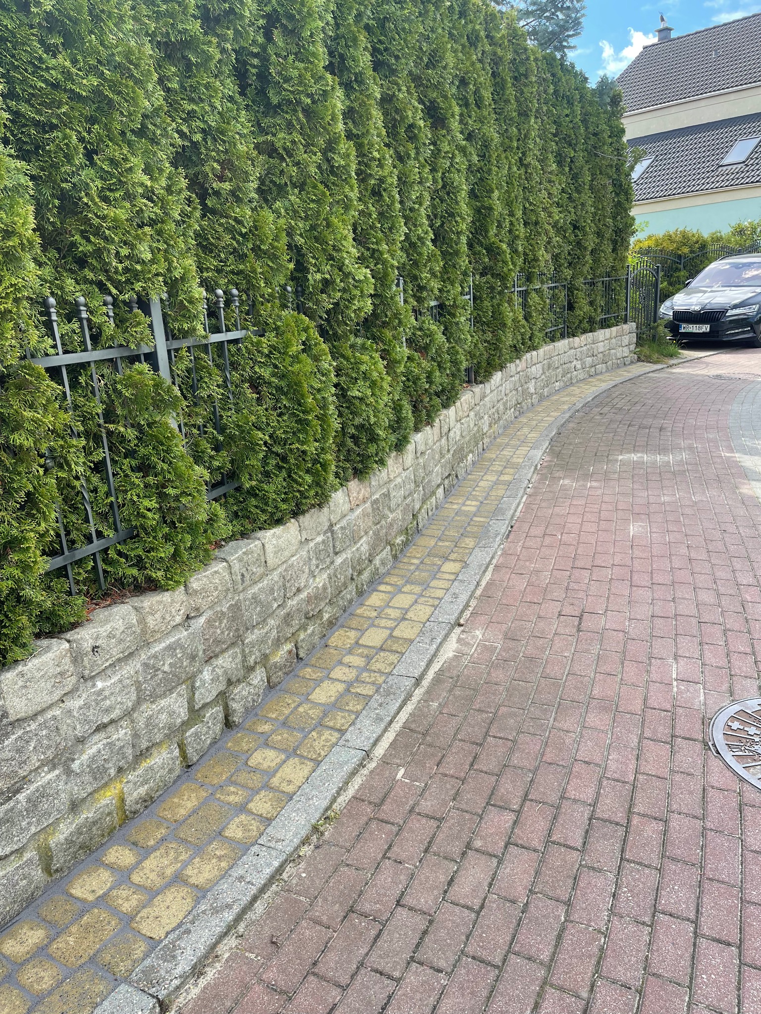Czysta kostka brukowa wzdłuż żywopłotu z tui, oddzielona kamiennym murkiem. Widoczny fragment ulicy i zaparkowany samochód. Ciemna brama.