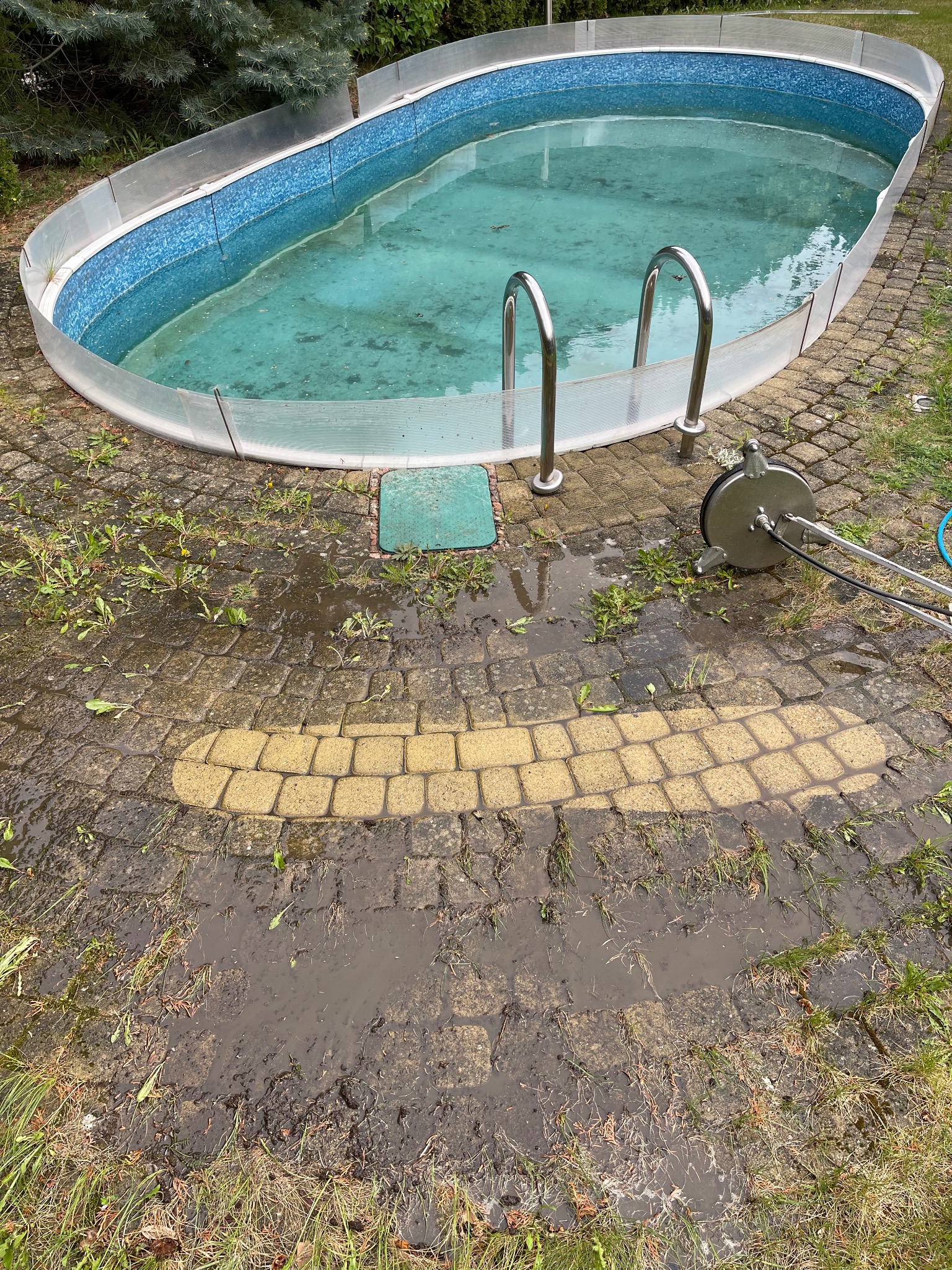 Czyszczenie kostki brukowej wokół basenu narzędziem wysokociśnieniowym, widoczny efekt czyszczenia, mokra powierzchnia, resztki roślinności.