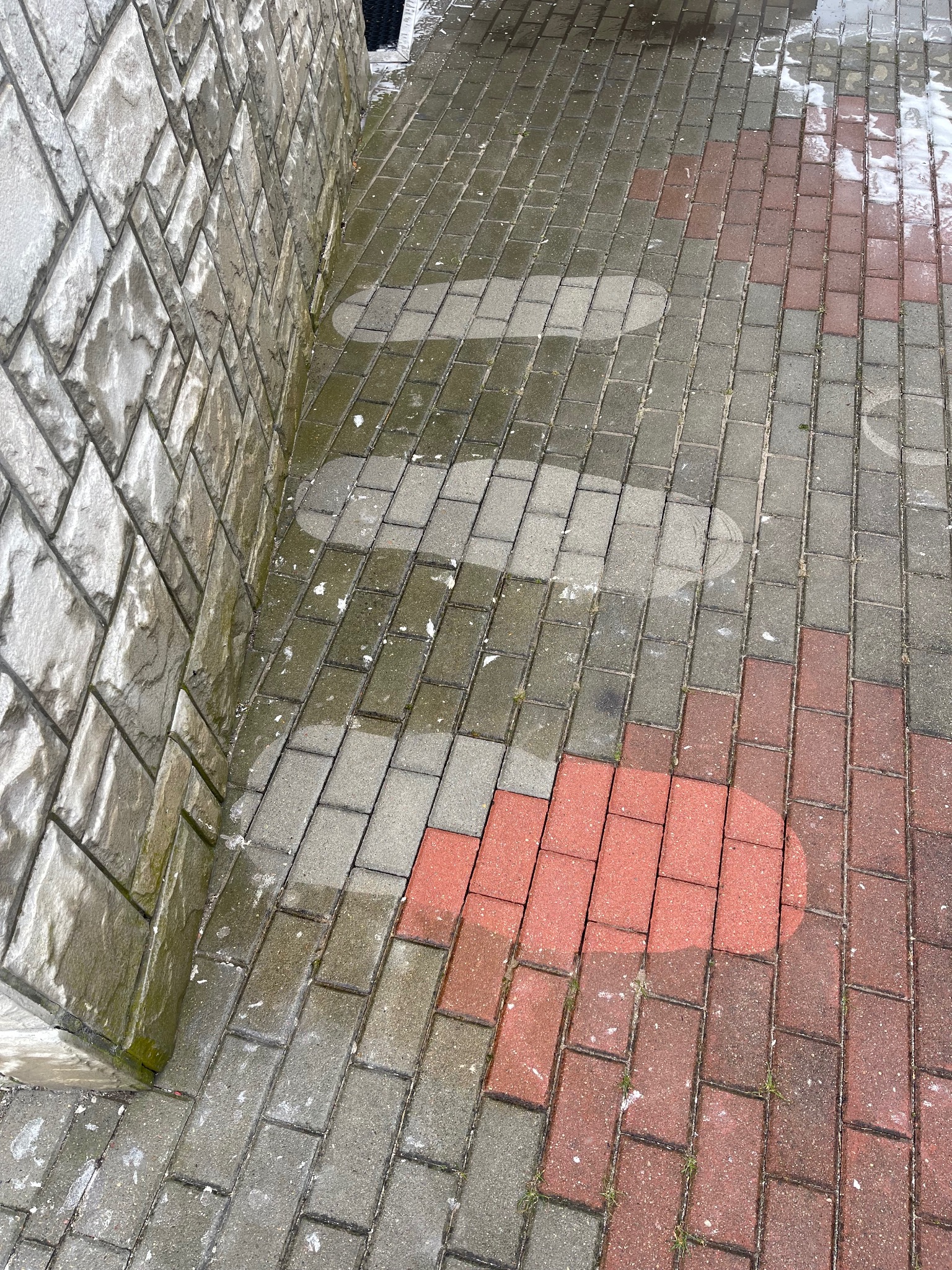 Mokry chodnik z kostki brukowej po czyszczeniu, z widocznymi jaśniejszymi, czystymi plamami, przylegający do kamiennej ściany budynku.