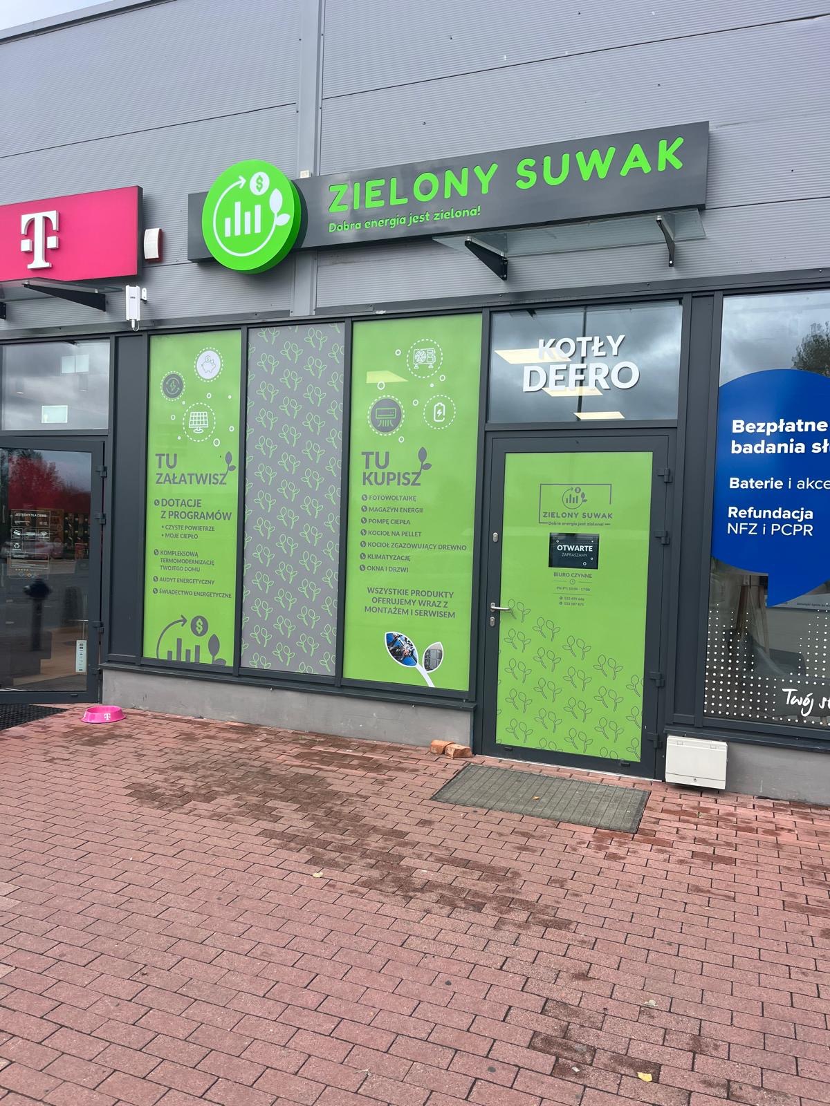 Fasada budynku firmy 'Zielony Suwak' z reklamami dotacji, fotowoltaiki, pomp ciepła, kotłów i okien. Widoczne logo firmy i informacje o ofercie.
