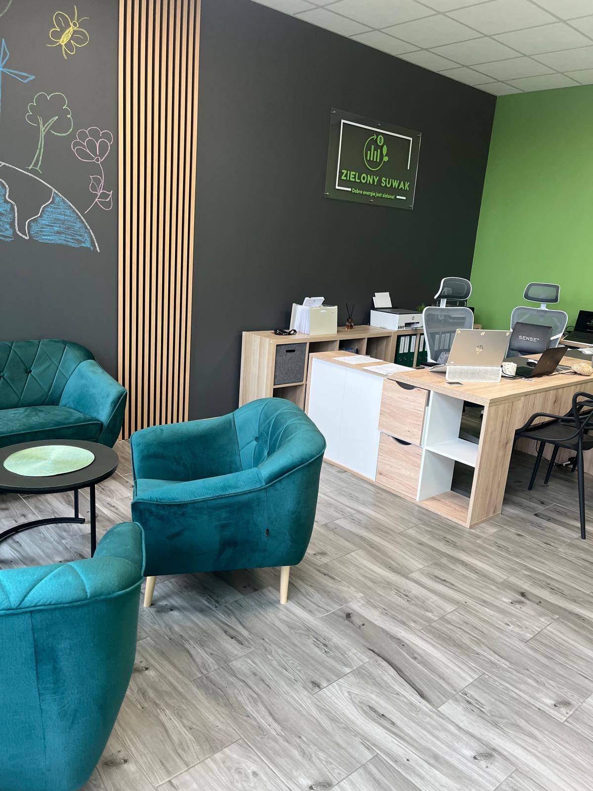 Nowoczesne biuro z zielonymi akcentami: turkusowe fotele, drewniane lamele na ścianie, logo 'Zielony Suwak' i biurko z laptopami. Jasne wnętrze z drewnianą podłogą.