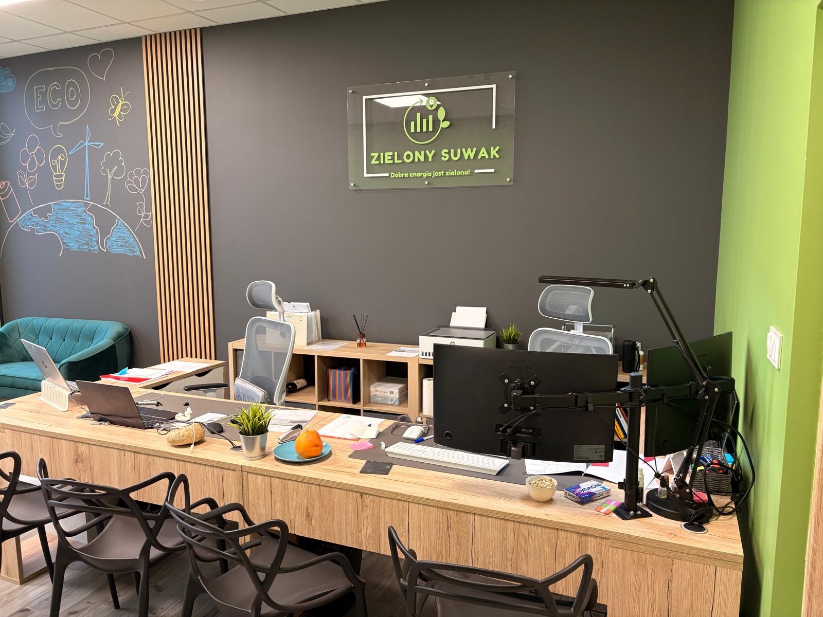 Nowoczesne biuro z drewnianym biurkiem, komputerami, krzesłami i ścianą z rysunkami ekologicznymi oraz logo firmy 'Zielony Suwak'.