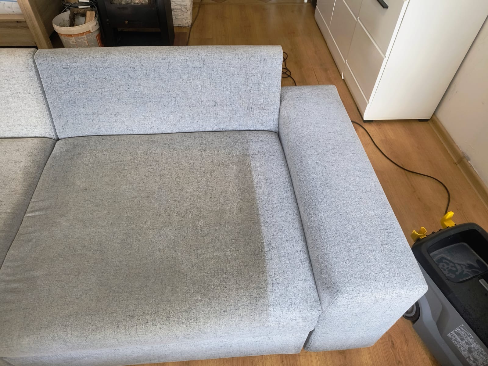 Jasnoszara kanapa z widoczną różnicą w czystości po praniu, z lewej strony widoczny brud, z prawej czysta tapicerka. W tle sprzęt do prania.