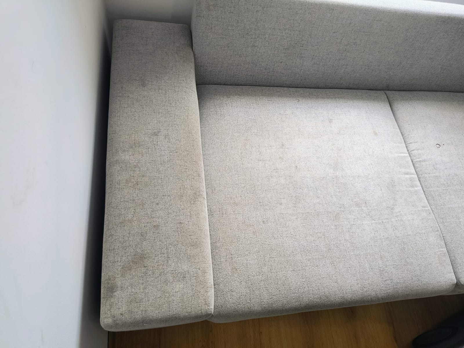 Szara, tekstylna kanapa z widocznymi plamami na oparciu i siedzisku, wymagająca profesjonalnego czyszczenia tapicerki. Widok z góry, naturalne oświetlenie.