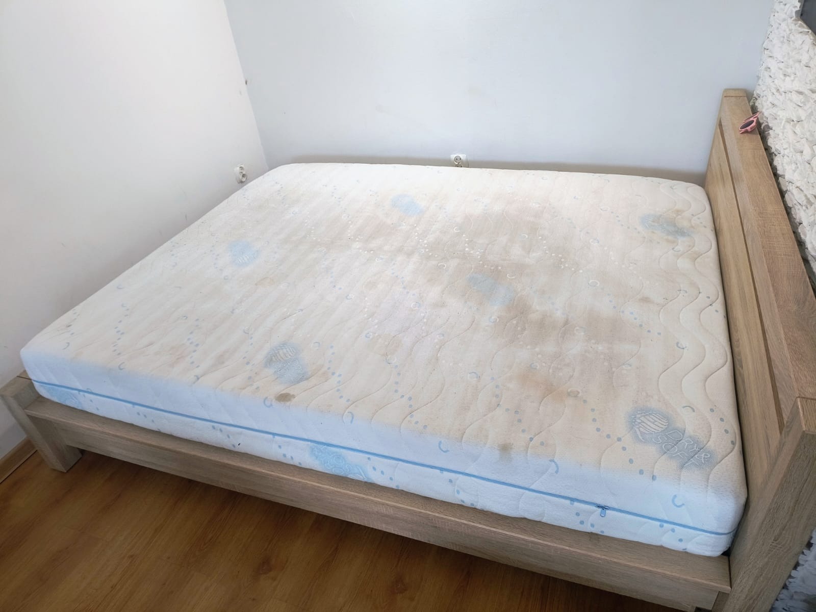 Materac na drewnianej ramie łóżka, z widocznymi plamami i zabrudzeniami na białym poszyciu. Perspektywa lekko z góry, ukazująca stan przed czyszczeniem.