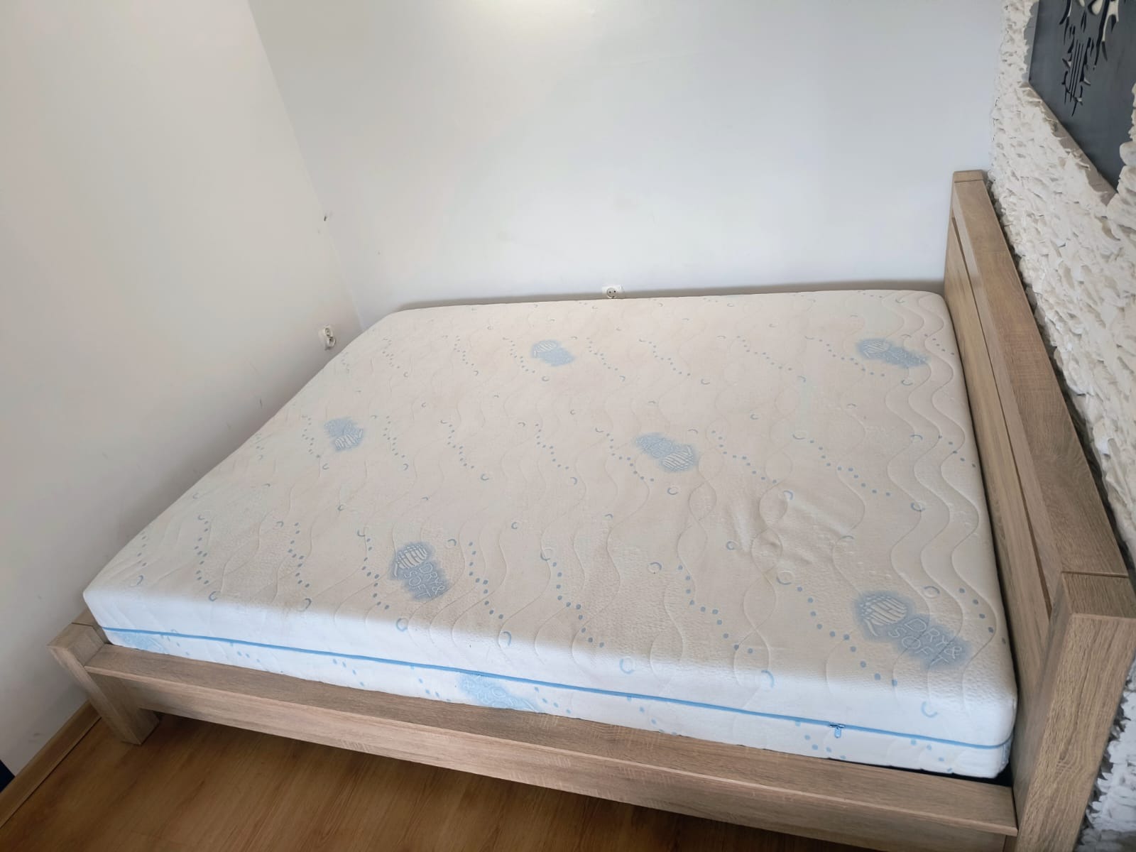 Materac na drewnianym łóżku w pokoju z białymi ścianami. Widoczne niebieskie wzory i zamek błyskawiczny na materacu. Ujęcie z perspektywy.