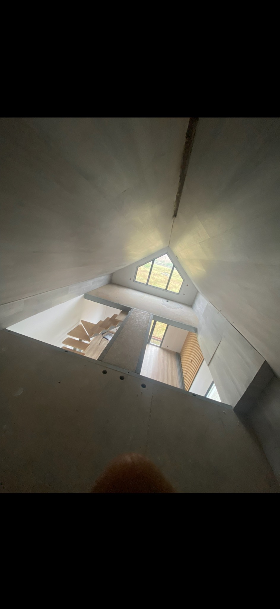 Wysoki pokój ze skosami, widok z dołu na drewniane schody i trójkątne okno. Ściany w szarym kolorze po szpachlowaniu, widoczne łączenia płyt.