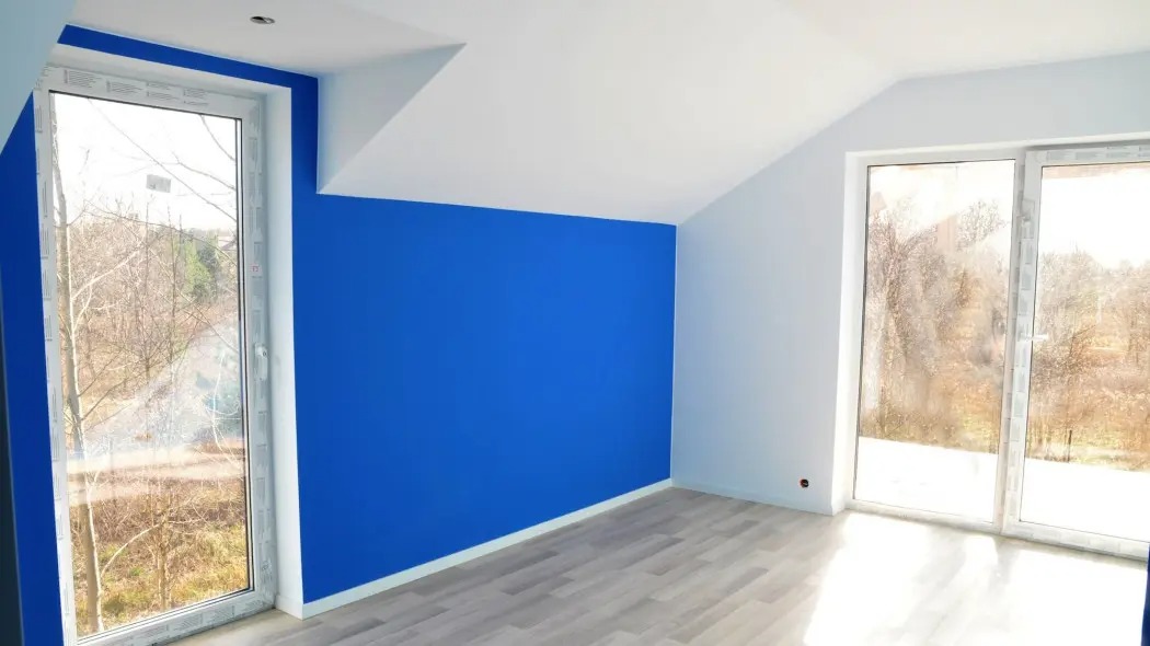 Błękitna ściana w pokoju na poddaszu z oknami i panelami. Świeżo pomalowane wnętrze z widokiem na drzewa za oknem. Kontrast bieli i intensywnego koloru.
