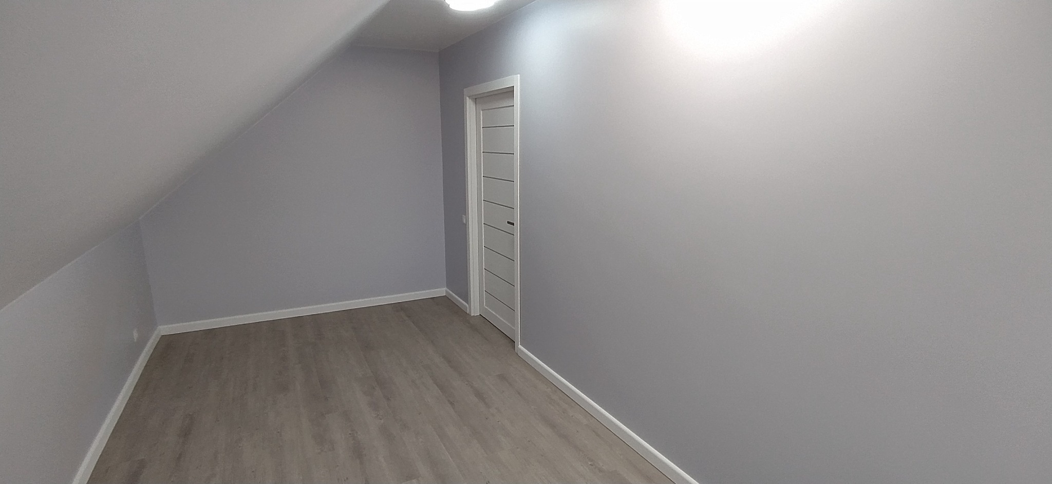 Poddasze po remoncie: świeżo pomalowane szare ściany, jasna podłoga z paneli i białe drzwi z poziomymi wgłębieniami. Skos dachu po lewej stronie.