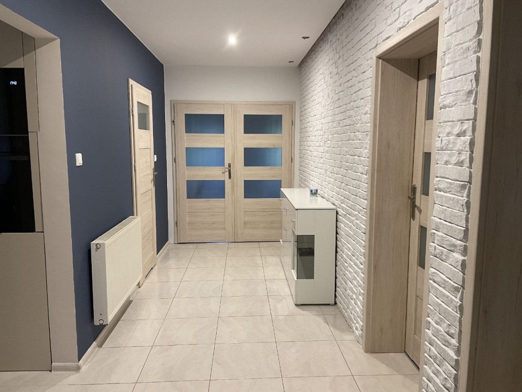 Korytarz po remoncie: biała cegła na ścianie, jasne panele podłogowe, dwoje drzwi z przeszkleniami i biała komoda. Granatowa ściana z grzejnikiem po lewej.