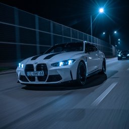 321AUTOBROKER.PL SPÓŁKA Z OGRANICZONĄ ODPOWIEDZIALNOŚCIĄ - Białe BMW M4 z czarnymi pasami na masce, jedzie nocą po oświetlonej drodze. W tle inne pojazdy i bariera dźwiękochłonna.