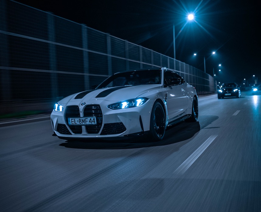 Białe BMW M4 z czarnymi pasami na masce, jedzie nocą po oświetlonej drodze. W tle inne pojazdy i bariera dźwiękochłonna.