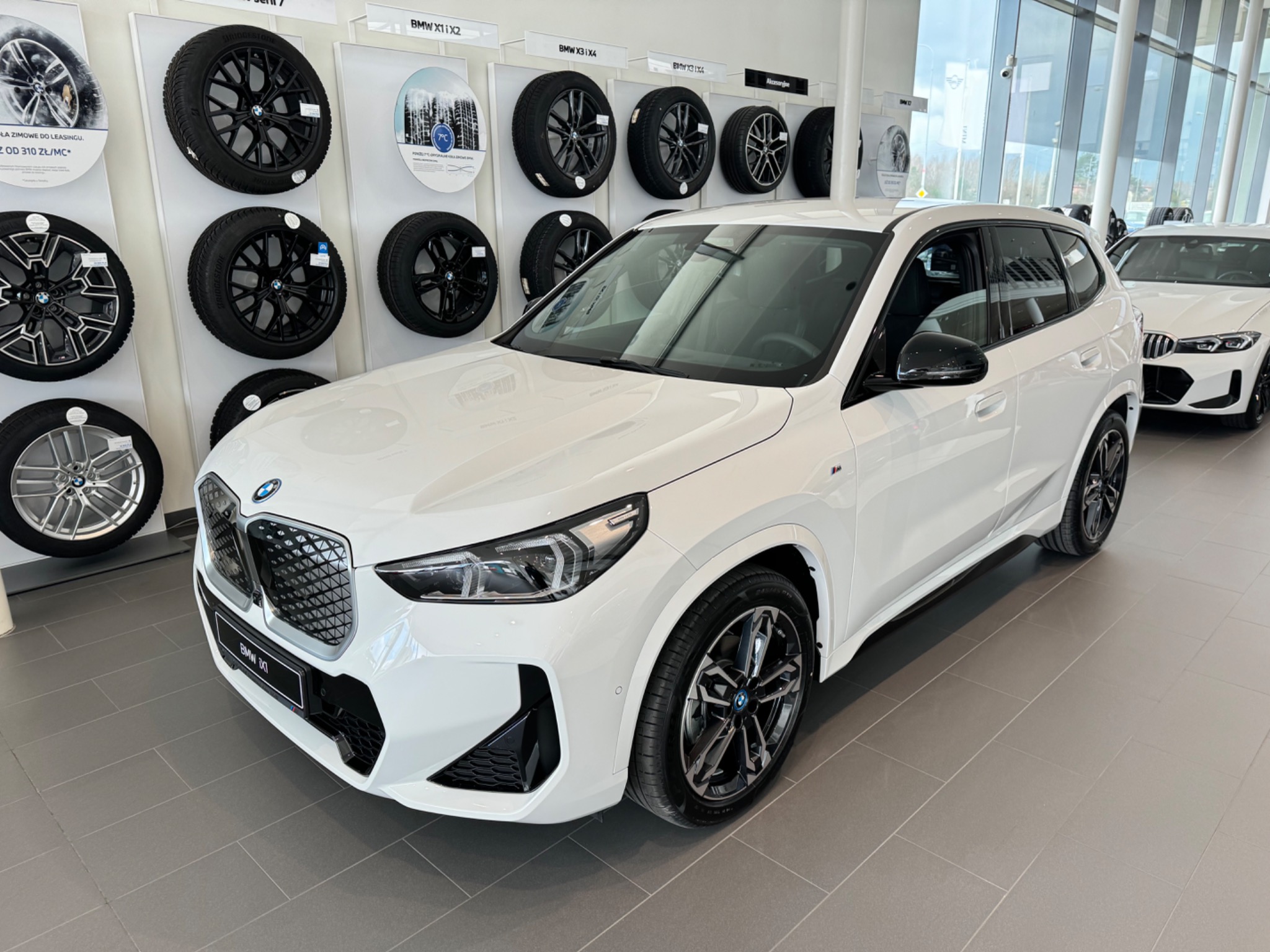 Nowy, biały BMW X1 w salonie samochodowym, z ekspozycją felg na ścianie w tle. Widoczne detale: czarne akcenty, felgi z logo BMW, panoramiczny dach.