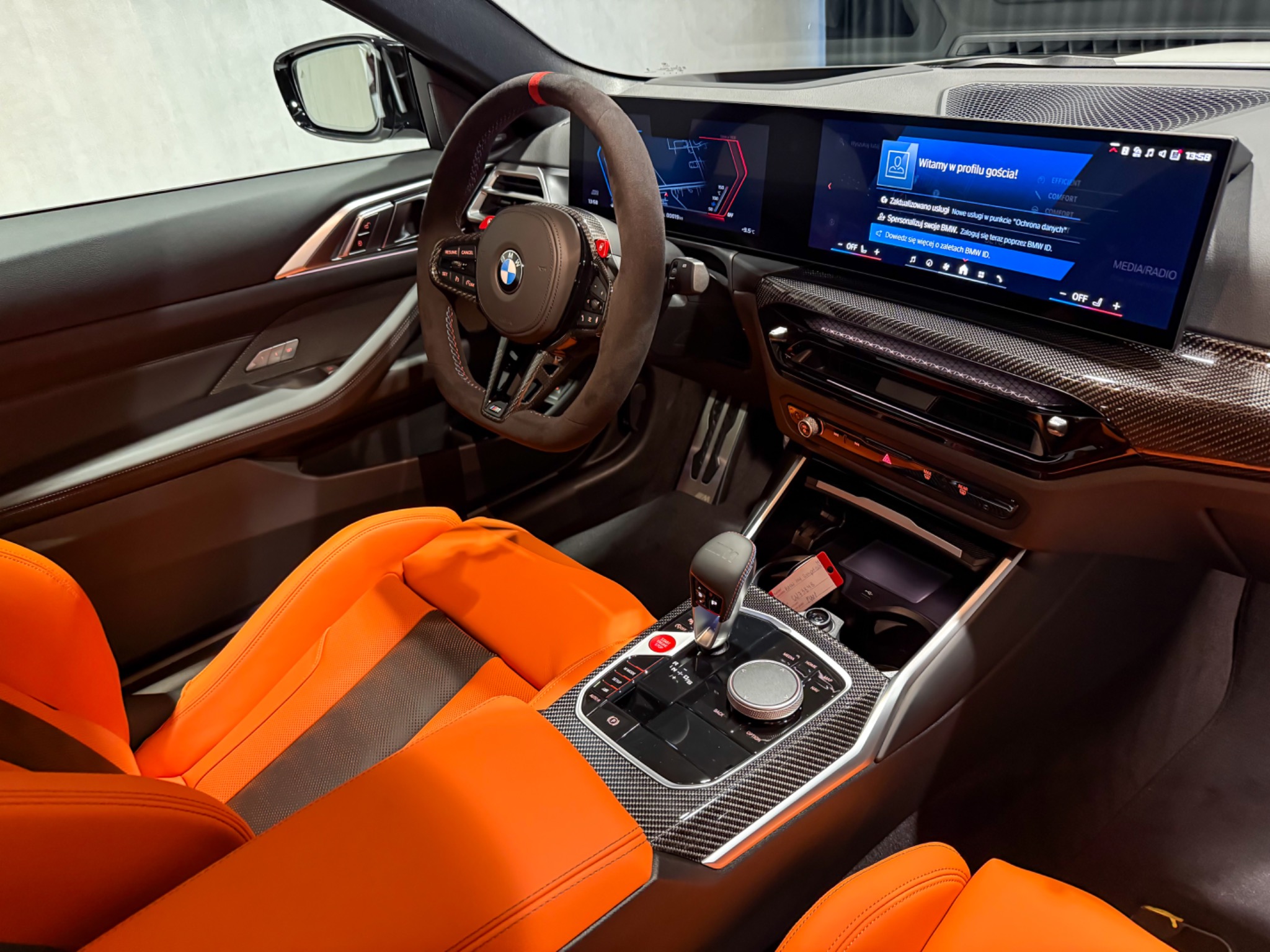 Wnętrze sportowego samochodu BMW z pomarańczowymi skórzanymi fotelami, karbonową konsolą i kierownicą obszytą alcantarą. Widoczny ekran systemu infotainment.